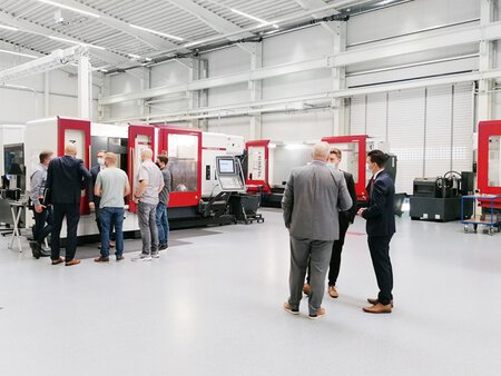 Besucher der Hausaustellung befinden isch im Technologiezentrum und schauen sich die ausgestellten HEDELIUS Bearbeitungszentren an. Zu sehen ist eine ACURA mit MARATHON Automation und im Hintergrund steht die TILTENTA 7