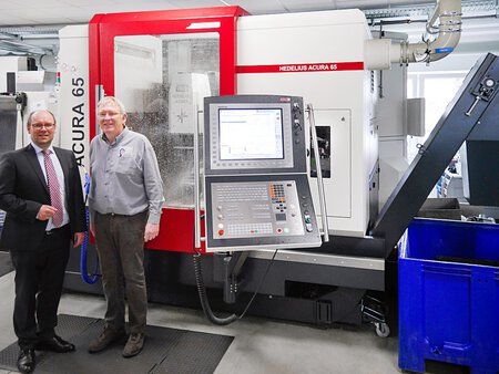 Blick auf das ACURA 65 5-Achs-Bearbeitungszentrum von HEDELIUS in der Fertigung der Pfisterer Feinwerktechnik GmbH am Chiemsee. Die Maschine ist in einer platzsparenden, kompakten Bauweise ausgeführt und befindet sich in einer modernen Werkhalle. Im Hintergrund sind Mitarbeiter bei der Arbeit zu sehen, die die hochpräzisen Bauteile für labor- und medizintechnische Geräte herstellen.