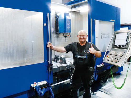 Aufnahme des 4-/5-Achs-Bearbeitungszentrums TILTENTA-7 in einer kundenindividuellen dunkelblauen RAL-Lackierung. Ein Anwender steht vor der Maschine und schaut zufrieden in die Kamera. Zu sehen ist der Innenraum der CNC-Fräse, der nach Kundenwunsch ausgestattet wurde.