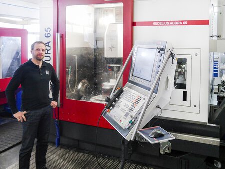 Dargestellt ist das HEDELIUS 5-Achs-Bearbeitungszentrum ACURA-65 in der Produktionshalle des Kunden. Die Maschine ist frontal abgebildet und unter Span. An der Maschine steht der Kunde mit Blick in die Kamera. Dank der kompakten Bauweise der ACURA-Baureihe findet sie bereits auf kleinstem Raum Platz und bietet dabei die volle Ausstattung und Automationsmöglichkeiten.