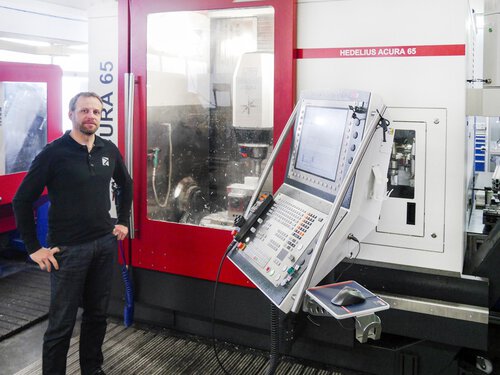 Dargestellt ist das HEDELIUS 5-Achs-Bearbeitungszentrum ACURA-65 in der Produktionshalle des Kunden. Die Maschine ist frontal abgebildet und unter Span. An der Maschine steht der Kunde mit Blick in die Kamera. Dank der kompakten Bauweise der ACURA-Baureihe findet sie bereits auf kleinstem Raum Platz und bietet dabei die volle Ausstattung und Automationsmöglichkeiten.
