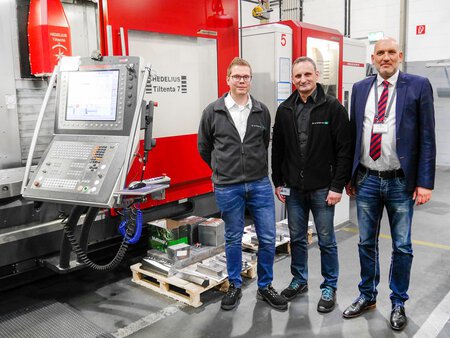 Bild des Hedelius 4-/5-Achs-Bearbeitungszentrums Tiltenta 7-2600, das für die präzise Zerspanung in der Fertigung eingesetzt wird. Die Maschine verfügt über einen Schwenkkopf, große Aufspannflächen und eine kompakte Bauweise, die eine flexible Bearbeitung verschiedener Werkstücke ermöglicht. Das Bild zeigt die technische Ausstattung und die Einsatzmöglichkeiten der Tiltenta 7-2600 in der industriellen Fertigung. An der Maschine stehen drei Anwender.